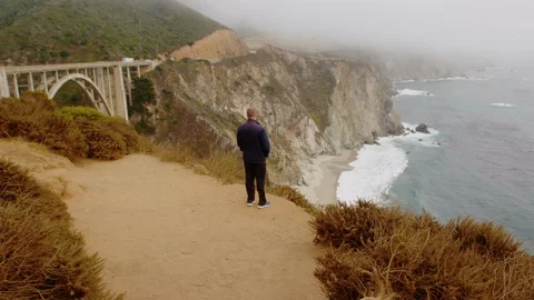 Lone man stands near dramatic cliffs at Big Sur, next to Bixby Creek Bridge Stock Footage 93755717