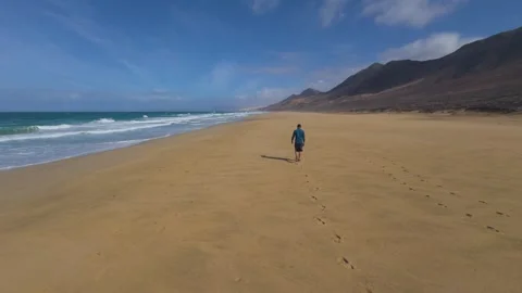 Lone man walking on empty beach coastline with ocean waves and mountains 스톡 동영상 331176043