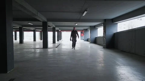 A lone man walks through a dimly lit, empty parking garage with concrete pillars Stock Footage 304444199