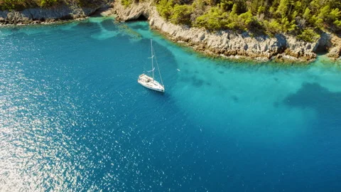 Lone sailboat floating in a bright blue cove surrounded by rocky forested cliffs Video stock 342771549