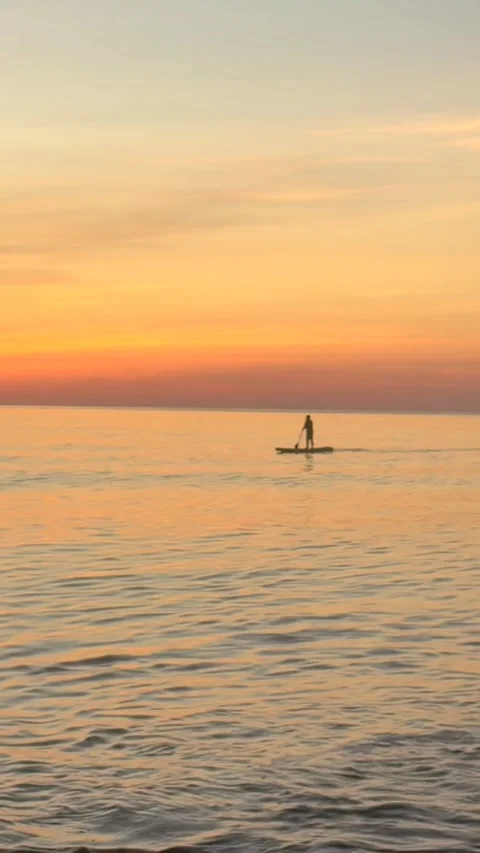 Lone stand up paddleboarder glides across calm sea at sunrise. Golden sky and Stock Footage 339897499
