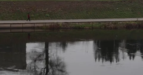 Lone walker by riverside path with mirrored silhouette on calm water Stock Footage 320167800