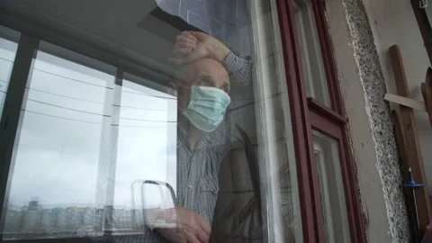 Lonely aged man in face mask looks through large window at home. Video stock 152572559