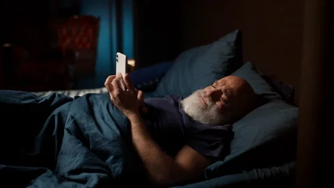 Lonely bearded man in his 50s scrolls on phone while lying awake with insomnia Stock Footage 342520899