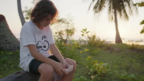 A lonely boy eats and bored against the background of the sunset, palm trees and Stock Footage 191451536