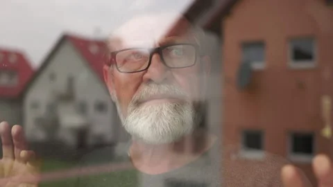 Lonely depressed gray-bearded elderly man with glasses stands by the window and Video stock 167691964