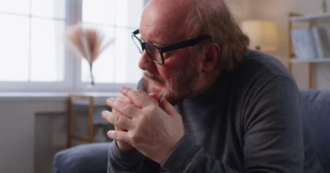 Lonely elderly man sitting on couch with clasped hands, looking down, feeling Stock Footage 337985244