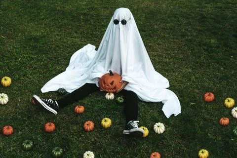 Lonely ghost sitting on a park surrounded by small pumpkins wearing glasses f Stock Photos
