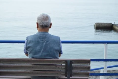 Lonely gray-haired elderly man sitting by the sea on a bench, the view from t Stock Photos