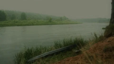 Lonely kayak lying in grass on the river bank in the rainy day near high trees Stock Footage 151109547