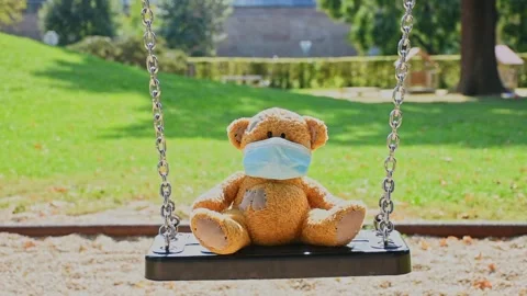 Lonely little teddy bear wearing a medical mask, standing on a swing in the p Stock Footage