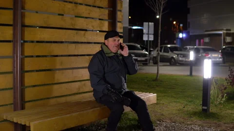 A lonely man in the evening sits on a bench and talks on the phone in cold Stock Footage 142269411