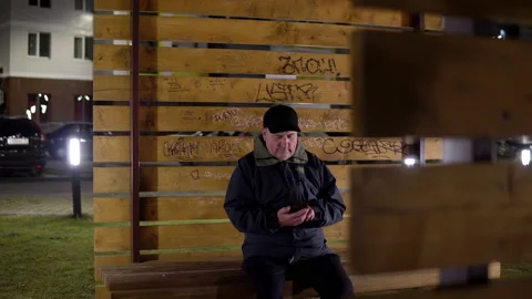 A lonely man in the evening sits on a bench and uses a smartphone in cold Video stock 142269964