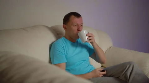Lonely man with a mug of tea sits on the sofa in the evening and watches TV Stock Footage 139545060