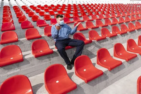 A lonely man with a protective mask on his face is sitting and watching a spo Stock Photos