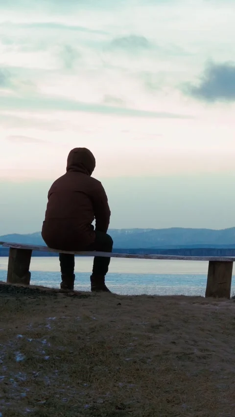 A lonely man sits timidly on a bench by a frozen lake at sunset. Stock Footage 296315309