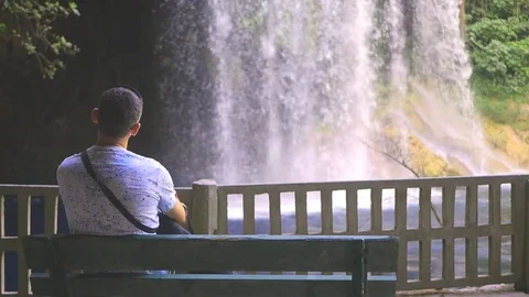 Lonely man sitting on a bench in the park with waterfall Stock Footage 77234298