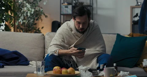 Lonely man is sitting on sofa, holding phone and scrolling. Boy is dressed in Stockbeeldmateriaal 218327837