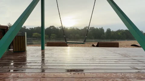 A lonely swing hangs over a wet wooden deck overlooking a calm lake Video stock 344979480