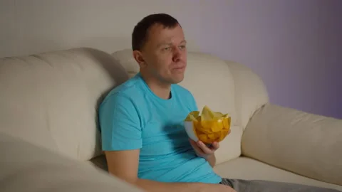 Lonely young man sits on the couch in the evening watching TV and eating chips Stock Footage 139286332
