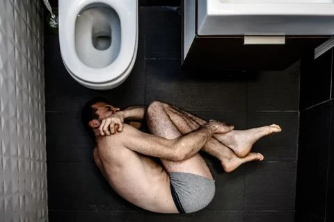 Lonely young man suffer inside in rest room alone. He lying on floor and holding Stock Photos