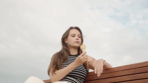 Lonely young woman eats ice cream while sitting on a bench under an overcast sky Stock-Footage 297209864