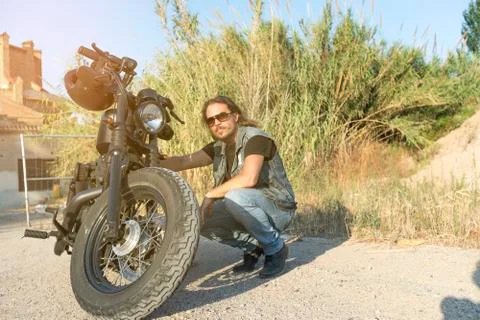 Long-haired brunette guy posing on a black custom motorcycle Stock Photos