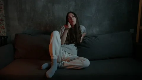 A long-haired brunette is sitting on the couch and switching channels to TV Stock Footage 197773277