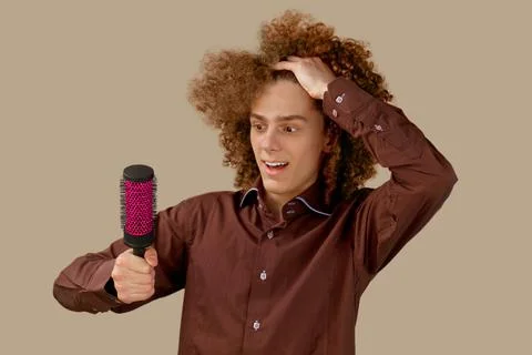 A long-haired curly-haired guy in a brown shirt on a beige background uses a  Stock Photos