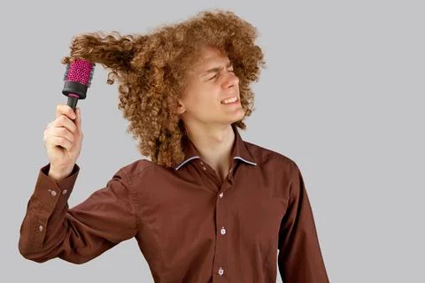 A long-haired curly-haired guy in a brown shirt on a gray background uses a m Stock Photos