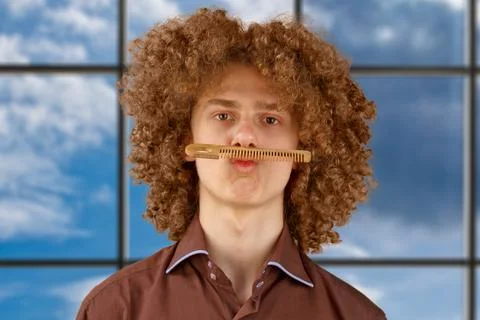 A long-haired curly-haired guy in a brown shirt on uses a wooden comb. Emotio Stock Photos