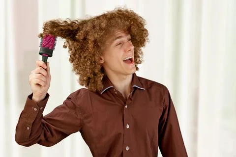 A long-haired curly-haired guy in a brown shirt uses a metal round comb. Emot Foto stock