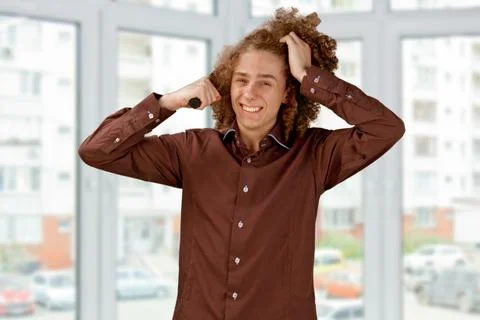 A long-haired curly-haired guy in a brown shirt uses a metal round comb. Emot Stock Photos