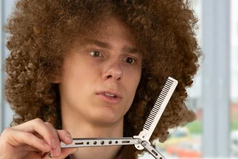 A long-haired curly-haired guy in a brown shirt uses a metal comb. Emotions b Stock Photos
