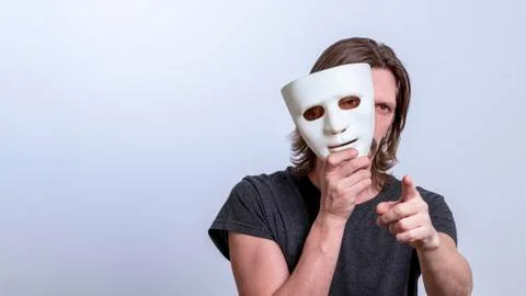 A long-haired man in a gray T-shirt looks out from behind a white mask and po Stock Photos