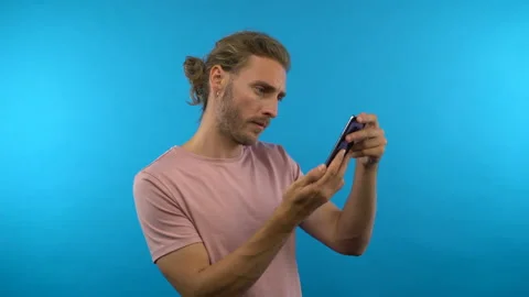 Long haired man on isolated blue background playing game on mobile phone 4K Stock Footage 138641771