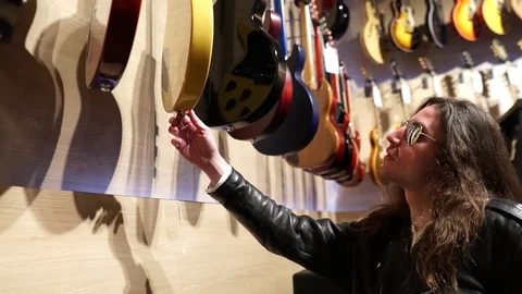 Long Haired Man with Leather Jacket checking selecting guitars in store Stock Footage 124671829