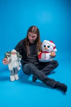 Long-haired man looking down at Santa's toy while holding a snowman and sitting Stock Photos