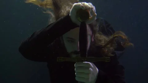 A long-haired man runs hand over the hilt of a sword in the dark water. close-up Vídeos de archivo 201142263