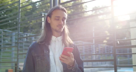 Long haired young guy chatting on smartphone while standing in sunlight near Stock-Footage 136647479