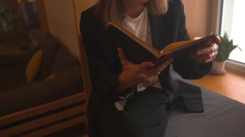 Long haired young man in black suit reads on windowsill aged book in black cover 스톡 동영상 198027118