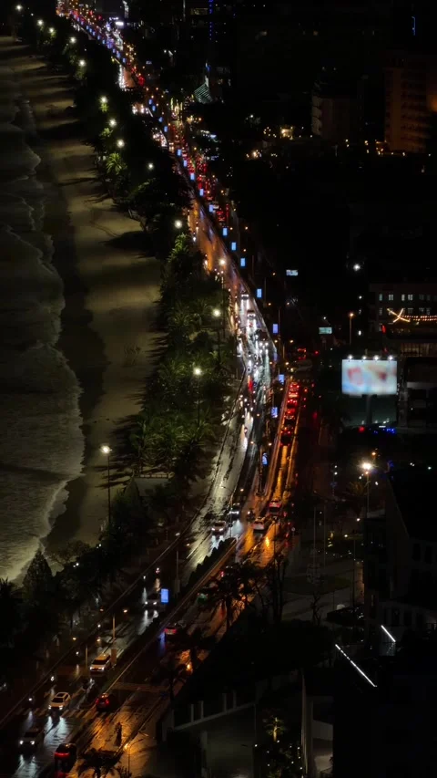 Long lines of headlights trace a coastal road at night as waves wash onto the Video stock 325514282
