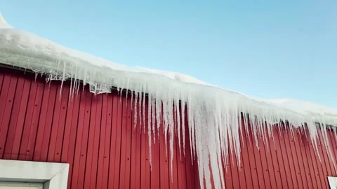 Long sharp icicles hanging from snow-covered roof edge of red barn against clear Stock Footage 341657338