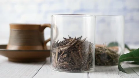 Loose leaf tea in clear glass jars sits on white wooden table with ceramic cu Stock Footage 332579694