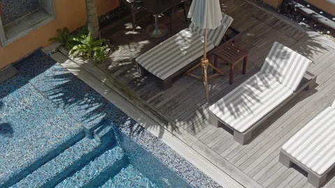 Lounge chairs side poolside in the shade of Palm tree on bright sunny day, Stock Footage 107404080
