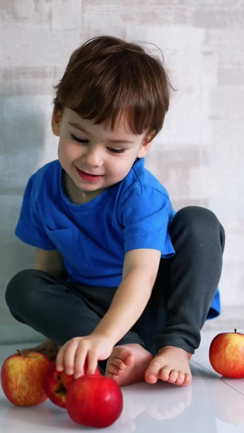 Lovely boy sitting at the wall playing with red apples. Kid puts the fruit from Stock Footage 342249228