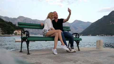 A loving couple doing selfie sitting on a bench near a mountain lake 스톡 동영상 92135852