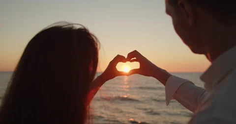 Loving couple make heart shape with hands in the sunlight at sunset on the beach Stock Footage 220730097