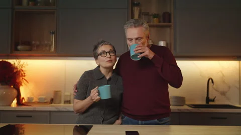 Loving couple in their 50s hugging and talking while drinking tea in kitchen Stock Footage 340920176