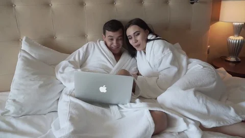 Loving couple in white coats on bed resting and having fun behind a macbook Stock Footage